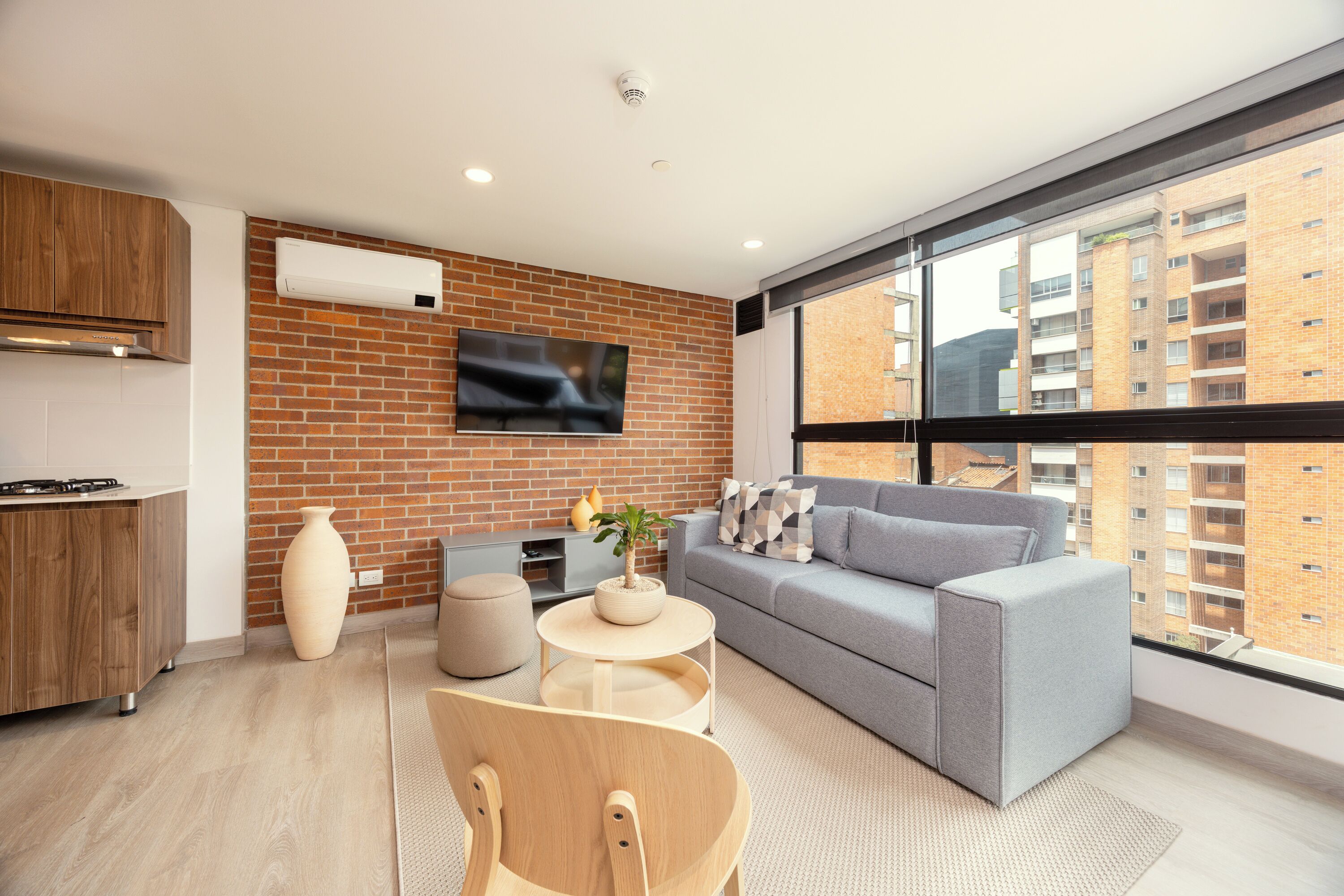 A modern apartment with a kitchen, a grey sofa, a TV mounted on a brick wall, and a large window overlooking a cityscape.