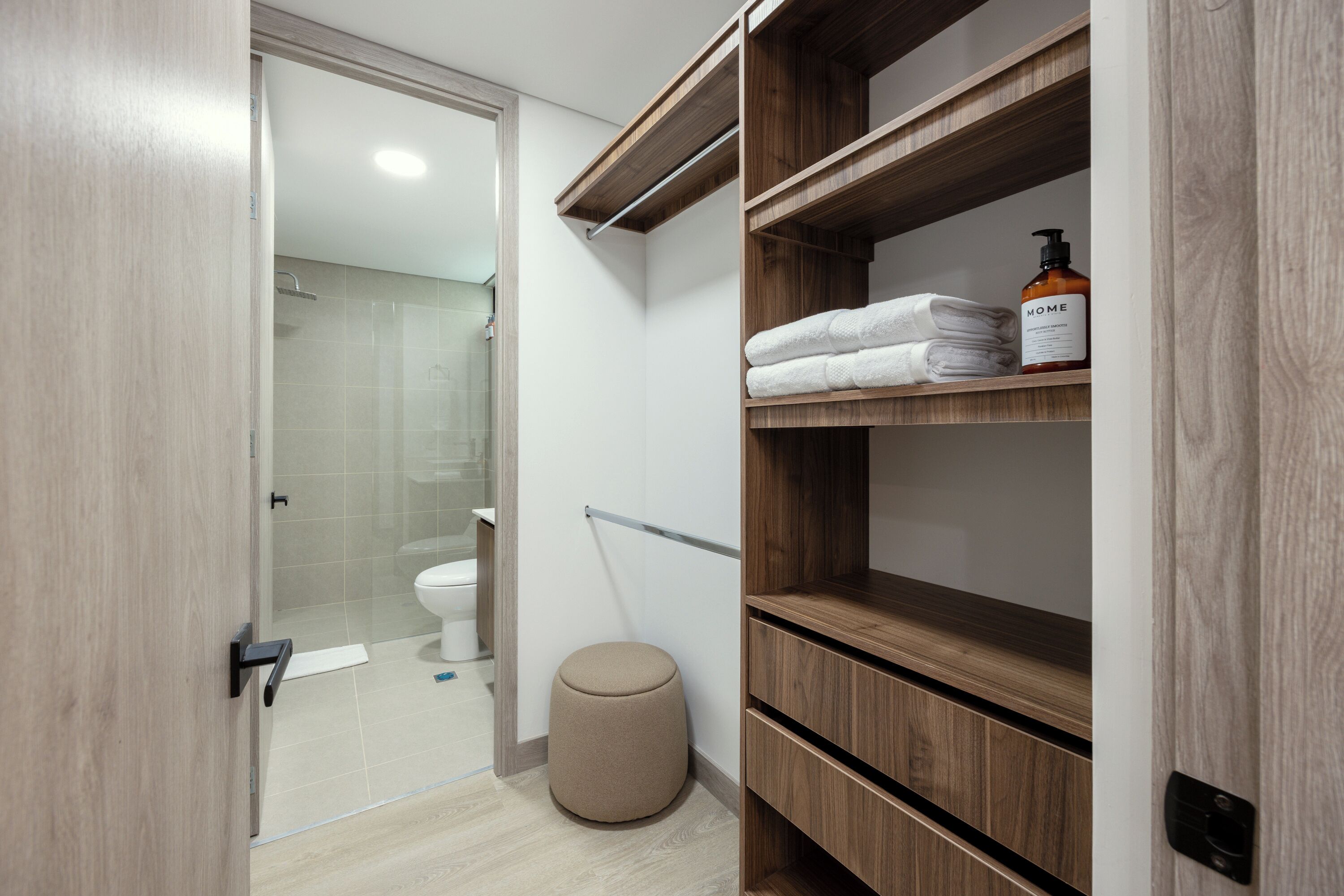 A modern bathroom with a glass shower enclosure, a wooden vanity with a towel rack, and a small round stool.