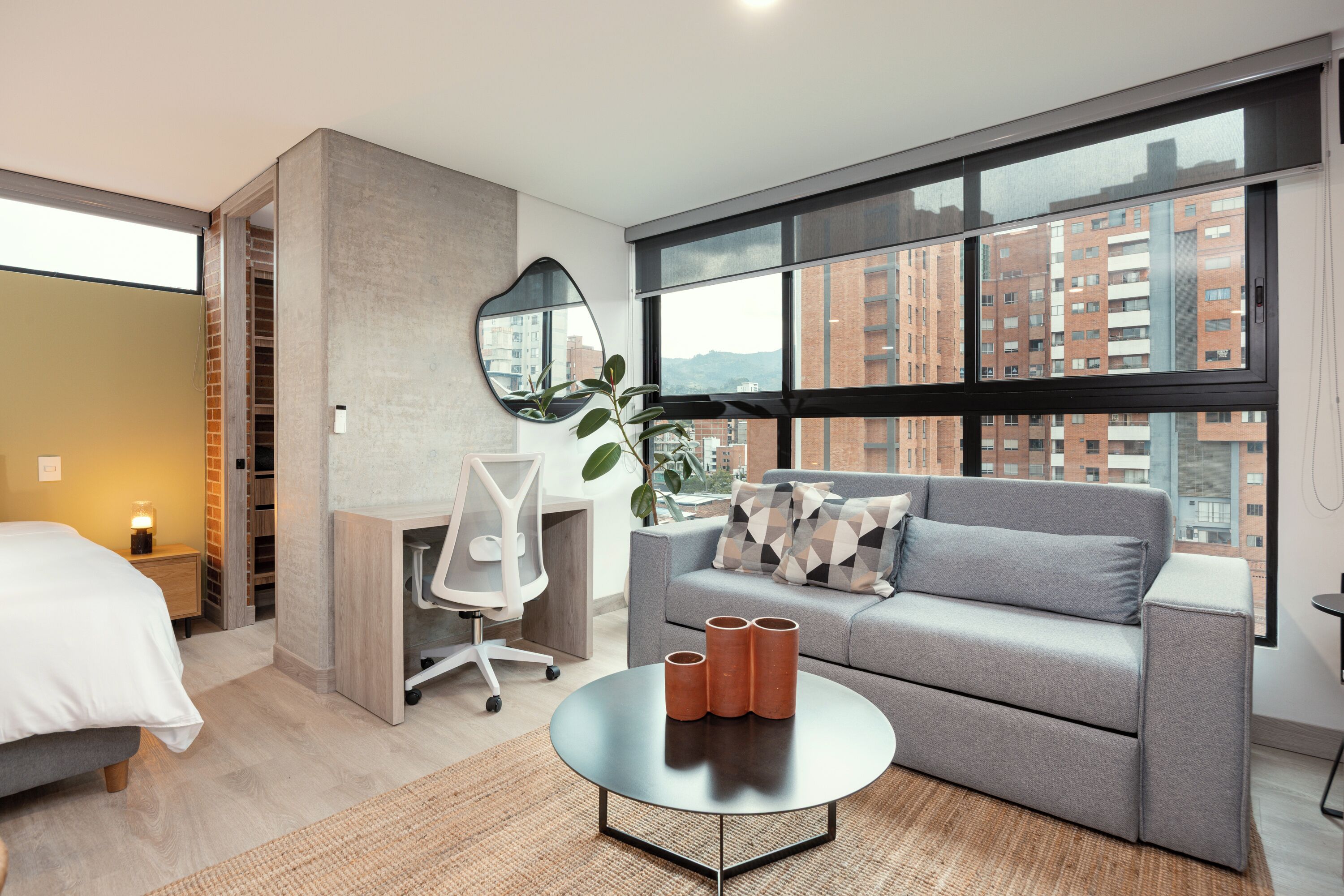 A modern hotel room with a grey sofa, a desk, a large window, and a view of buildings.