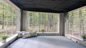 A cozy reading nook with a large window overlooking a forest, a cushioned bench, and a small plant on the windowsill. - Little Can in the Pines - Bunkie No. 1 (SIMCOE)