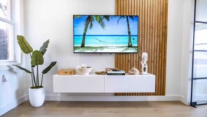 Una sala moderna con un televisor de pantalla plana montado en una pared de paneles de madera, una mesa consola blanca con objetos decorativos y una planta grande en maceta.