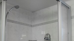 Une douche fermée par des parois en verre, dotée d’un pommeau de douche à main, d’un siège intégré et d’une petite étagère.