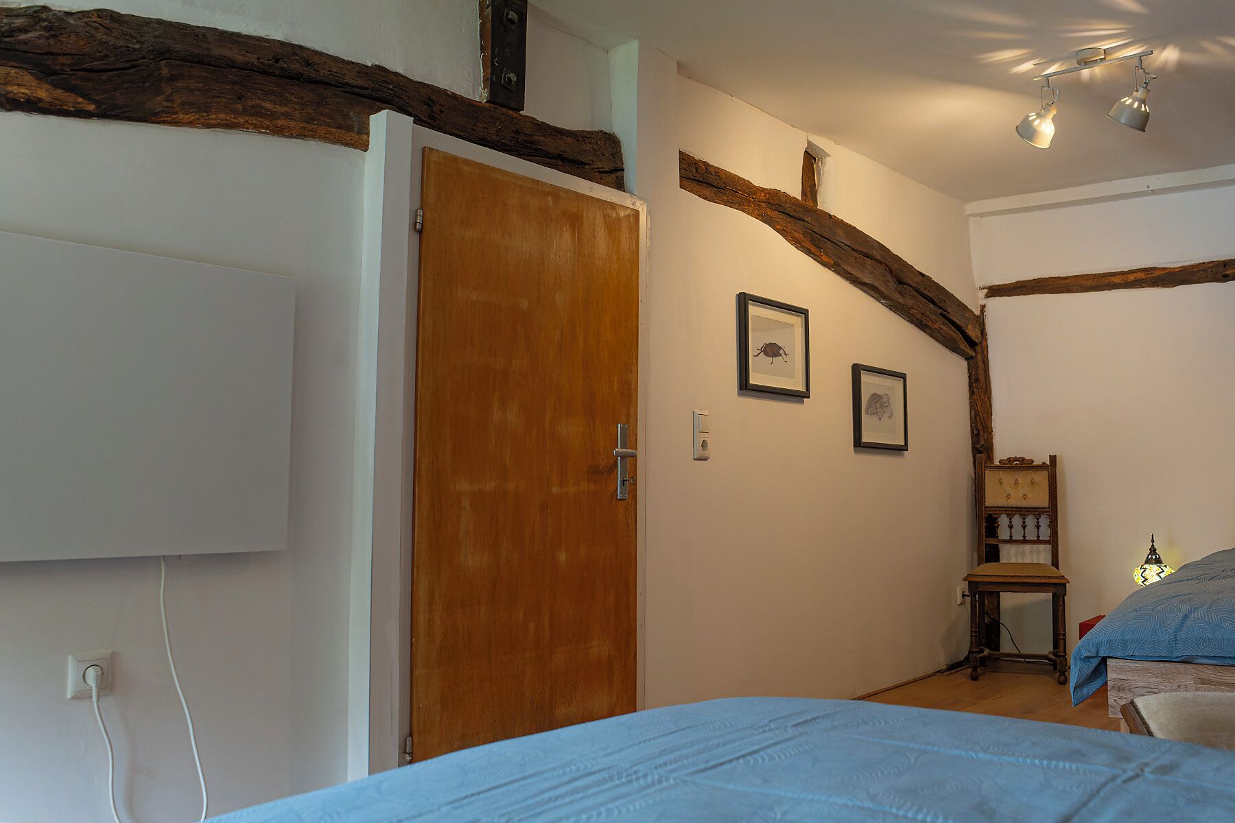 A bedroom with a wooden door, a bed with a blue bedspread, a small wooden chair, and a wall-mounted shelf.