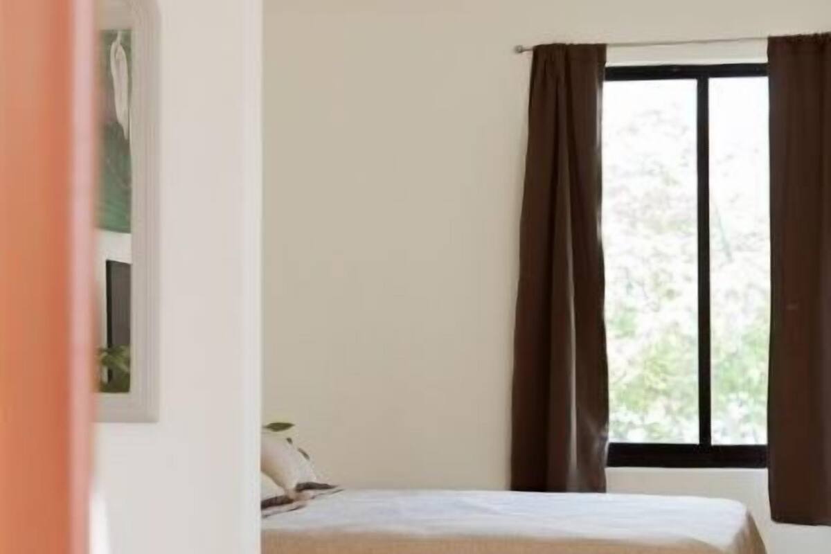 A bedroom with a bed, a window with curtains, a small table, and a wall-mounted shelf.