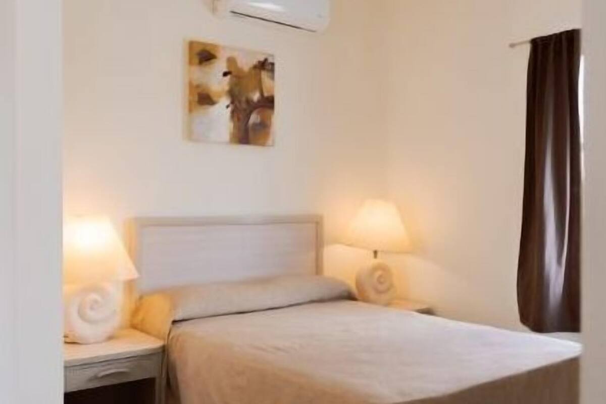 A hotel room with a bed, two bedside lamps, an air conditioning unit, and a wall-mounted artwork.