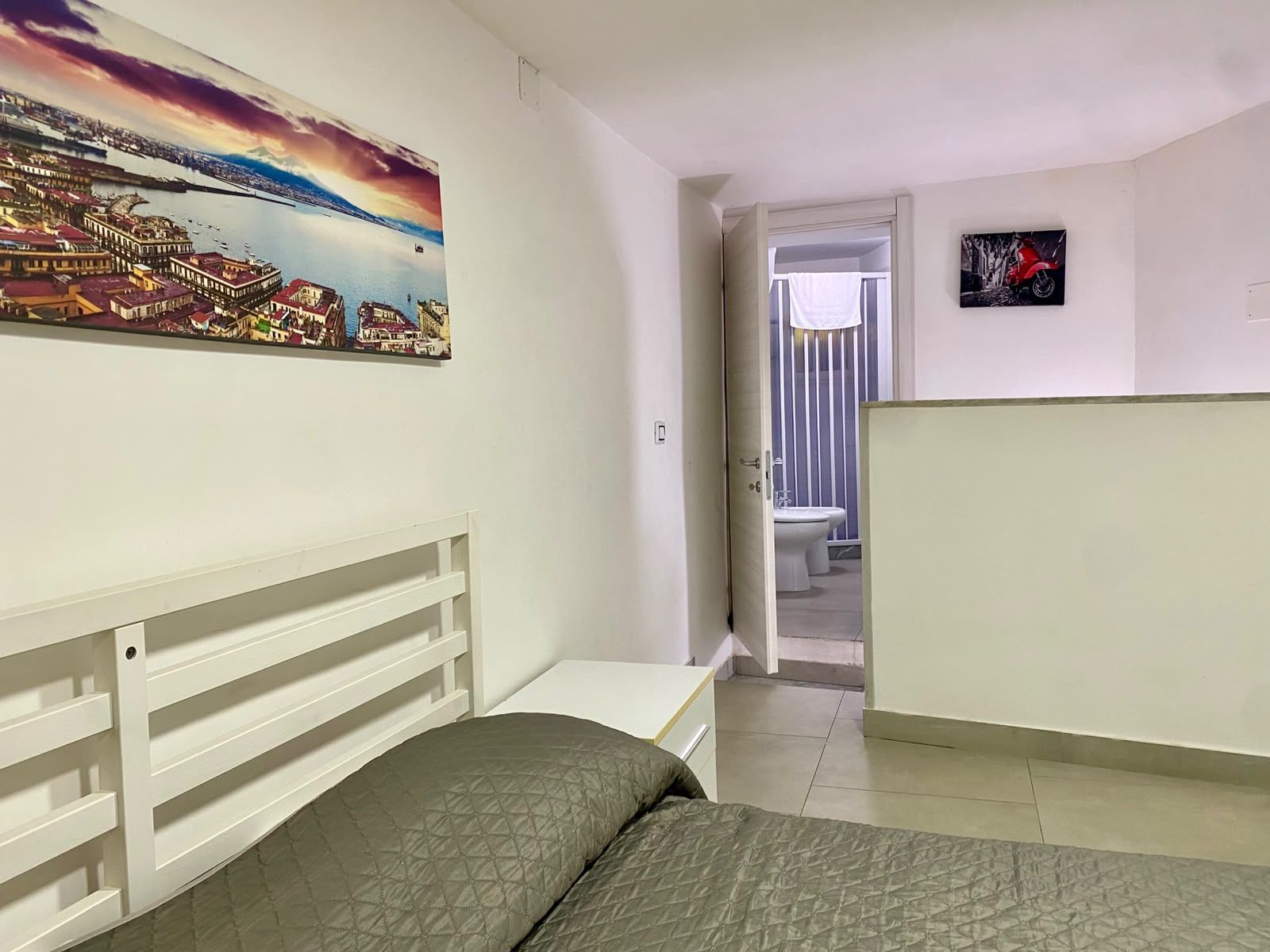 Een slaapkamer met een bed, een schilderij van een stadsgezicht, een badkamer met toilet en wastafel, en een deur naar een andere kamer.