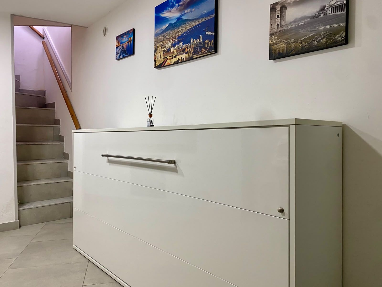 Un pasillo con una escalera, un armario blanco con pomo y tres cuadros en la pared.