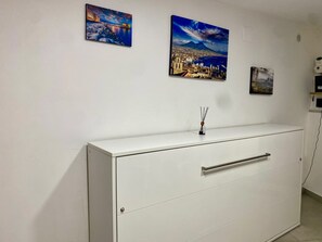 Un meuble blanc avec des tiroirs, un petit flacon de diffuseur de parfum à bâtonnets et deux tableaux encadrés accrochés au mur.