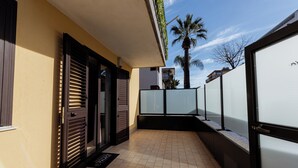 A corridor with a tiled floor, a door with a window, and a palm tree in the background. - APARTMENT 1 (Misterbianco)