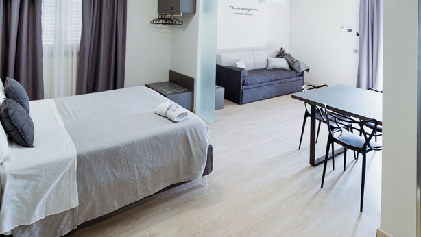 A modern hotel room with a bed, a desk, and a chair. There is a small table and a couch in the corner. - APARTMENT 2 (Misterbianco)