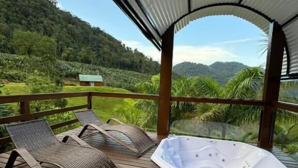A wooden deck with two lounge chairs and a hot tub, overlooking a lush green landscape and hills. - Recanto Fogo na Taça (Jaraguá do Sul)