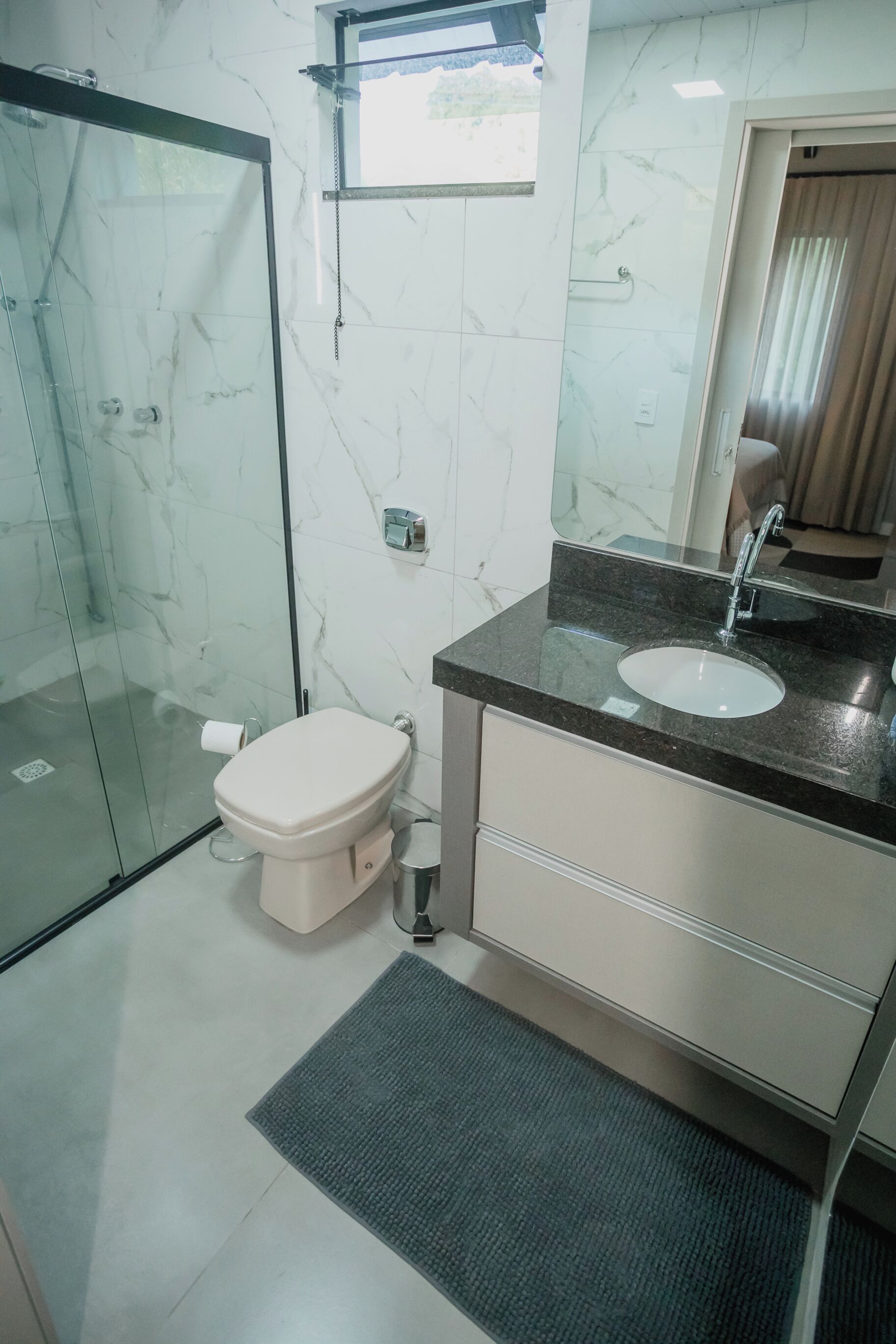 A modern bathroom with a glass shower enclosure, a white toilet, a sink with a mirror, and a small window.