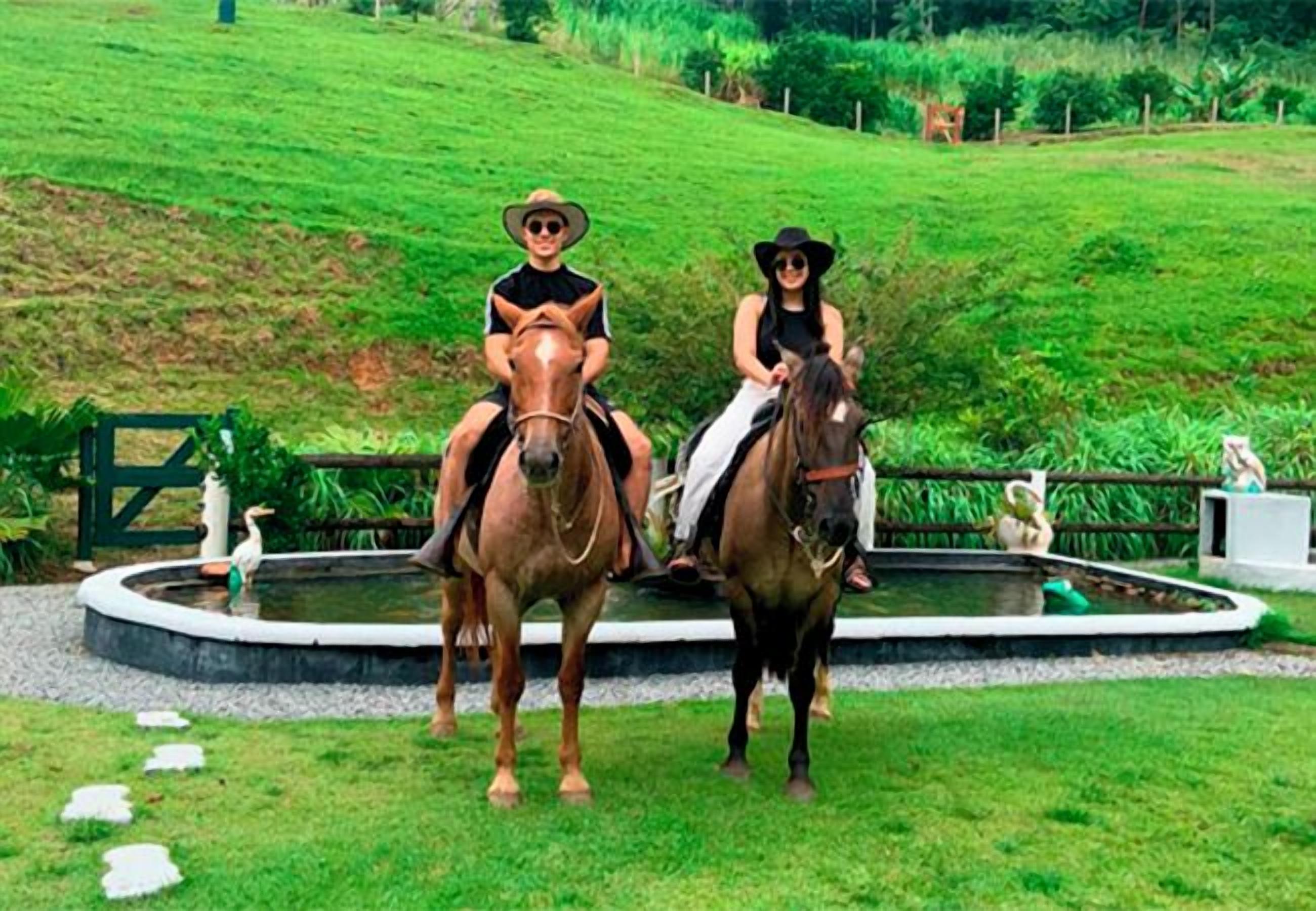 Dos personas montando a caballo en un parque con un estanque y una colina cubierta de hierba al fondo.
