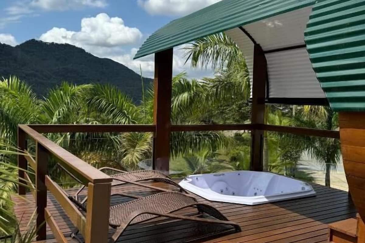 A wooden deck with a hot tub and lounge chairs, surrounded by palm trees and a mountain backdrop.