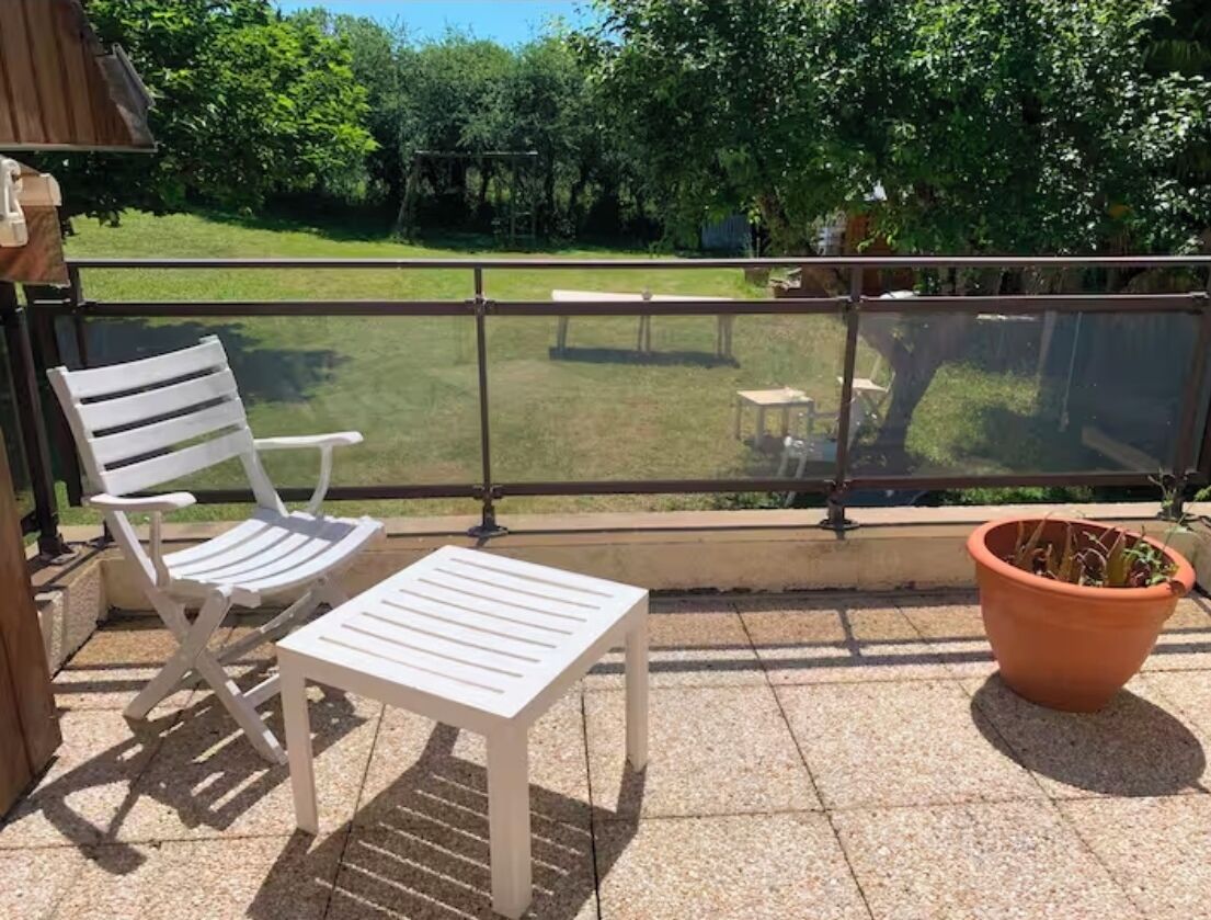 A balcony with a white chair and table, a potted plant, and a view of a grassy area with a table and chairs.