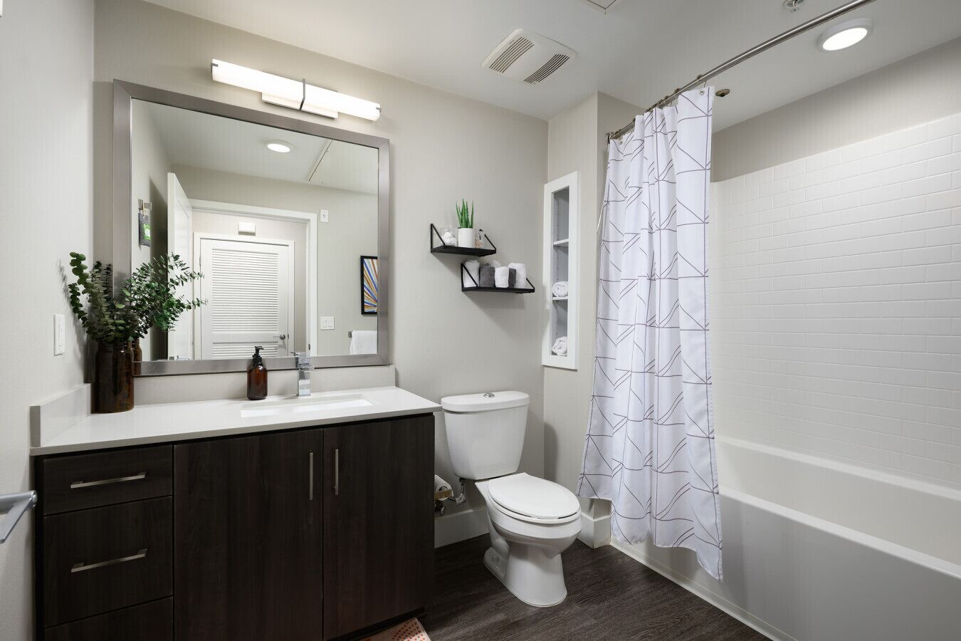 Une salle de bain moderne avec un meuble-lavabo blanc, des armoires en bois foncé, des toilettes blanches et un rideau de douche à motifs géométriques.