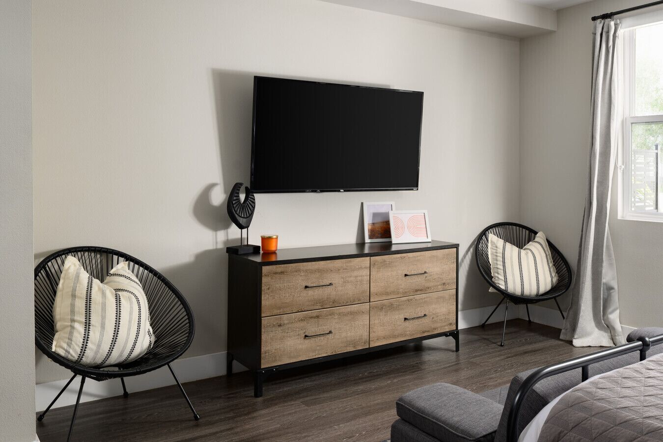 Ein modernes Schlafzimmer mit einem an der Wand befestigten Flachbildfernseher, einer Holzkommode, zwei schwarzen Stuhl mit Kissen und einem Bett mit grauer Steudecke.