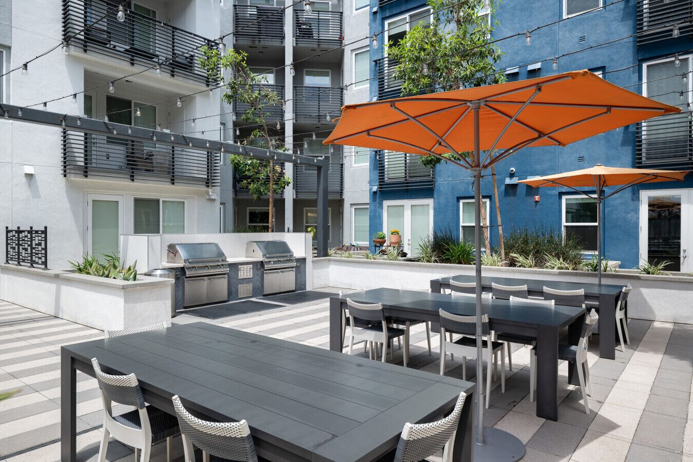Une terrasse extérieure équipée d’un barbecue, de tables et de chaises, entourée d’un immeuble bleu doté de balcons.