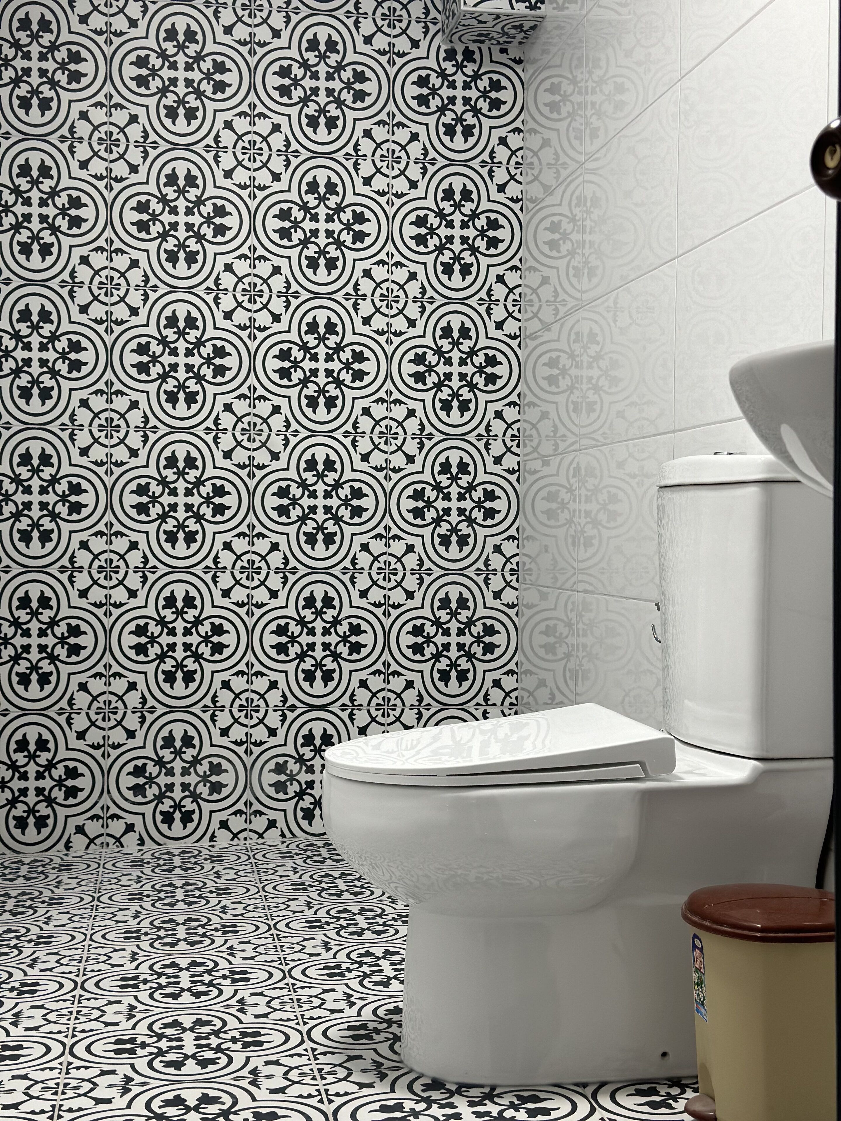 Une salle de bain avec un mur et un sol recouverts de carreaux à motifs, des toilettes et une poubelle.