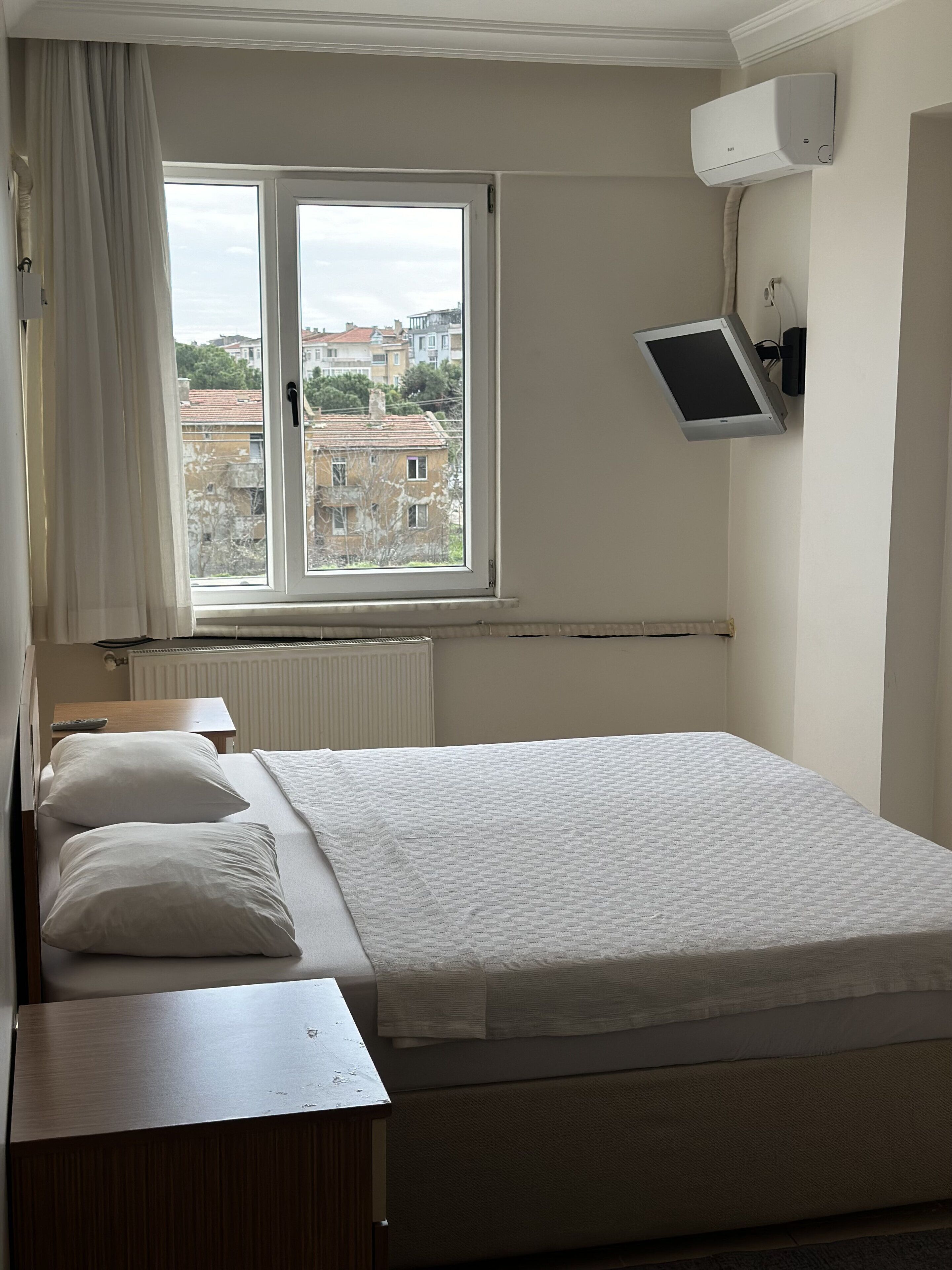 Una habitación de hotel con cama, mesita de noche, televisor montado en la pared y ventana con vistas a edificios.