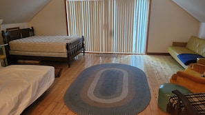 A bedroom with a bed, a sofa, a green ottoman, and a large window with blinds.