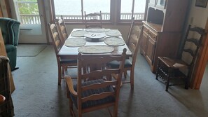 Un coin repas avec une table et des chaises en bois, une vue sur l’eau grâce à de grandes fenêtres, et un téléviseur posé sur un meuble en bois.