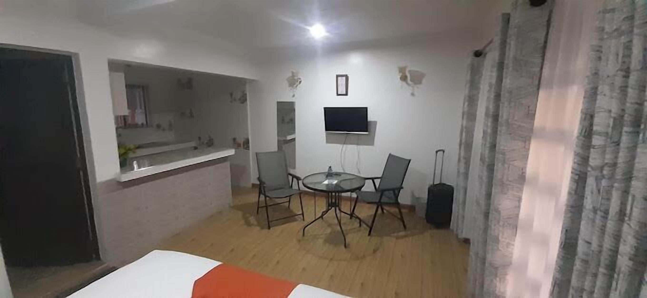 A compact living space with a kitchenette, a dining area with a round table and four chairs, a flat-screen TV mounted on the wall, and a curtain on the right side.