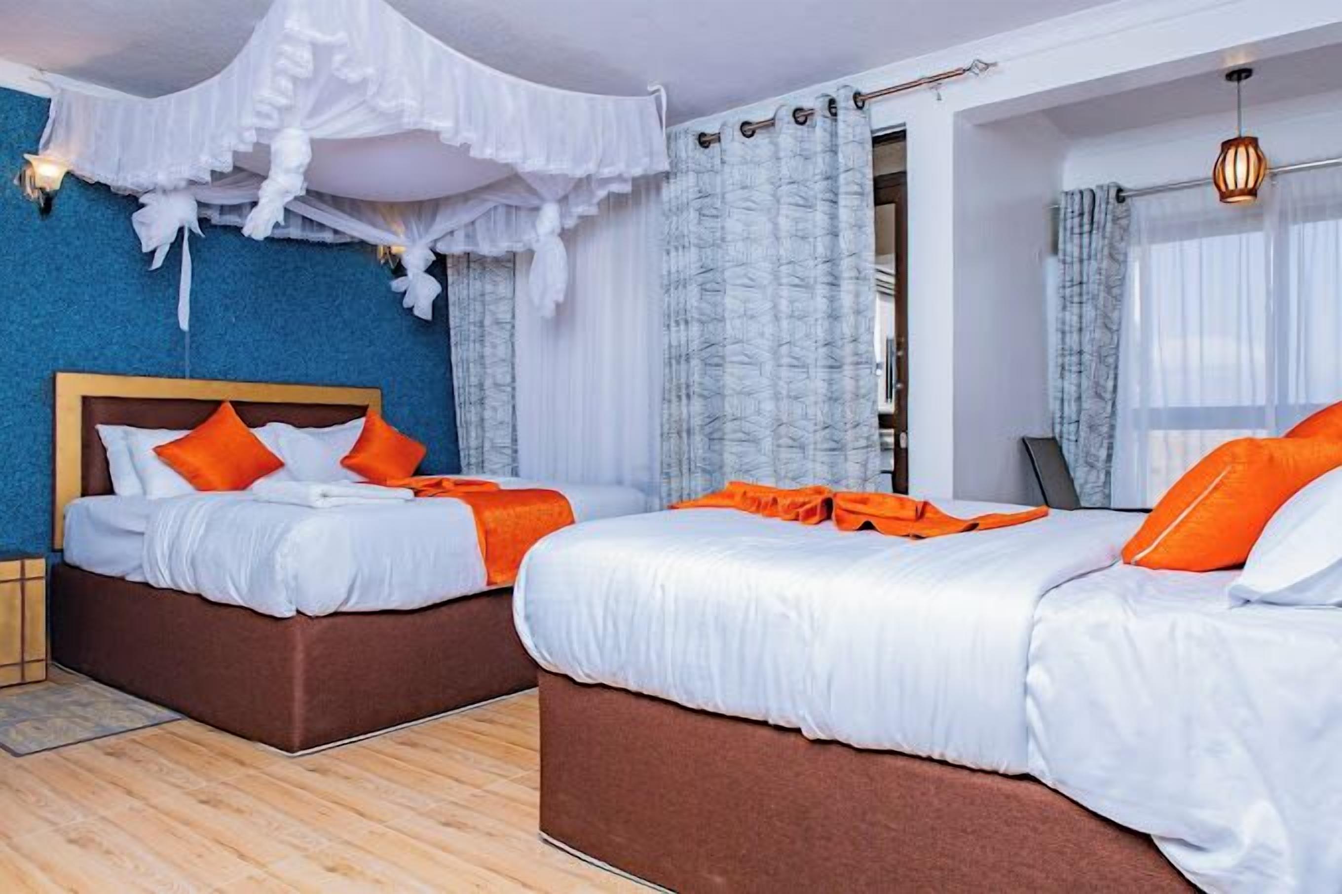 A hotel room with two beds, white bedding, orange pillows, a wooden headboard, a blue accent wall, and a window with curtains.