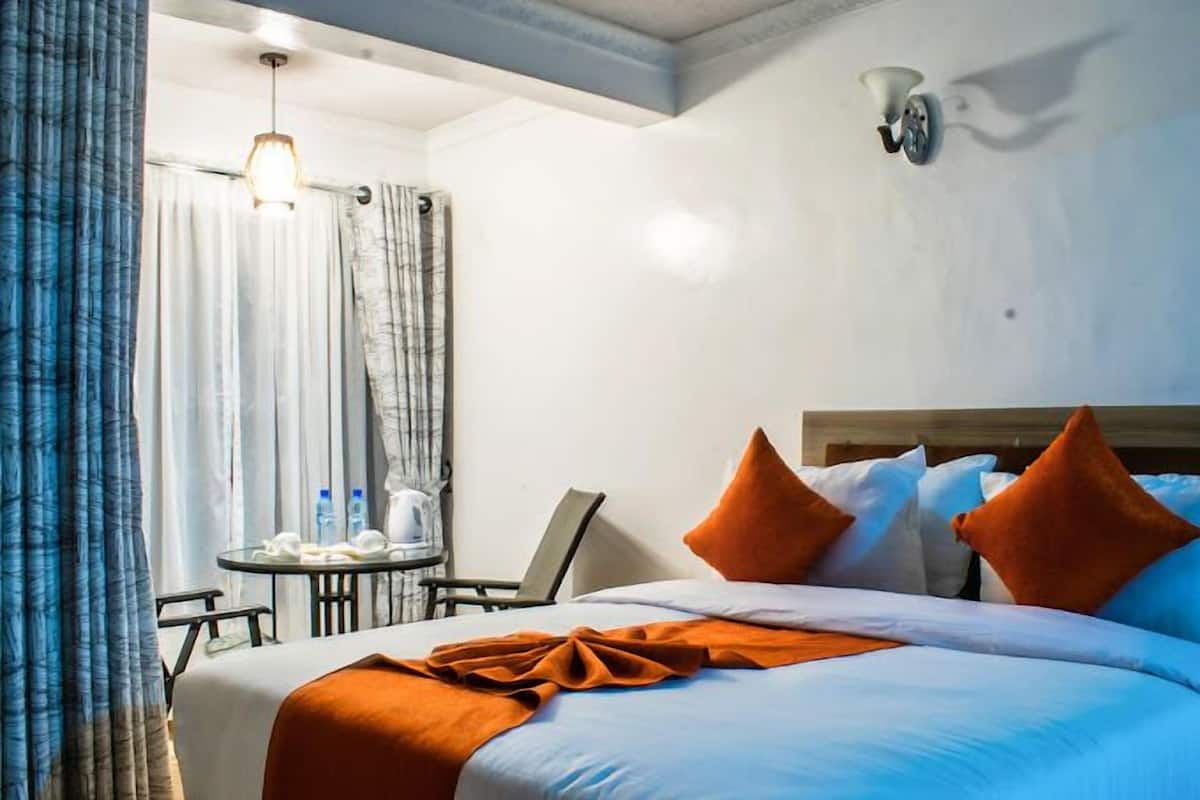 A hotel room with a bed, a small table with a chair, a lamp, and a window with curtains.