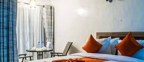 A hotel room with a bed, a small table with a chair, a lamp, and a window with curtains.