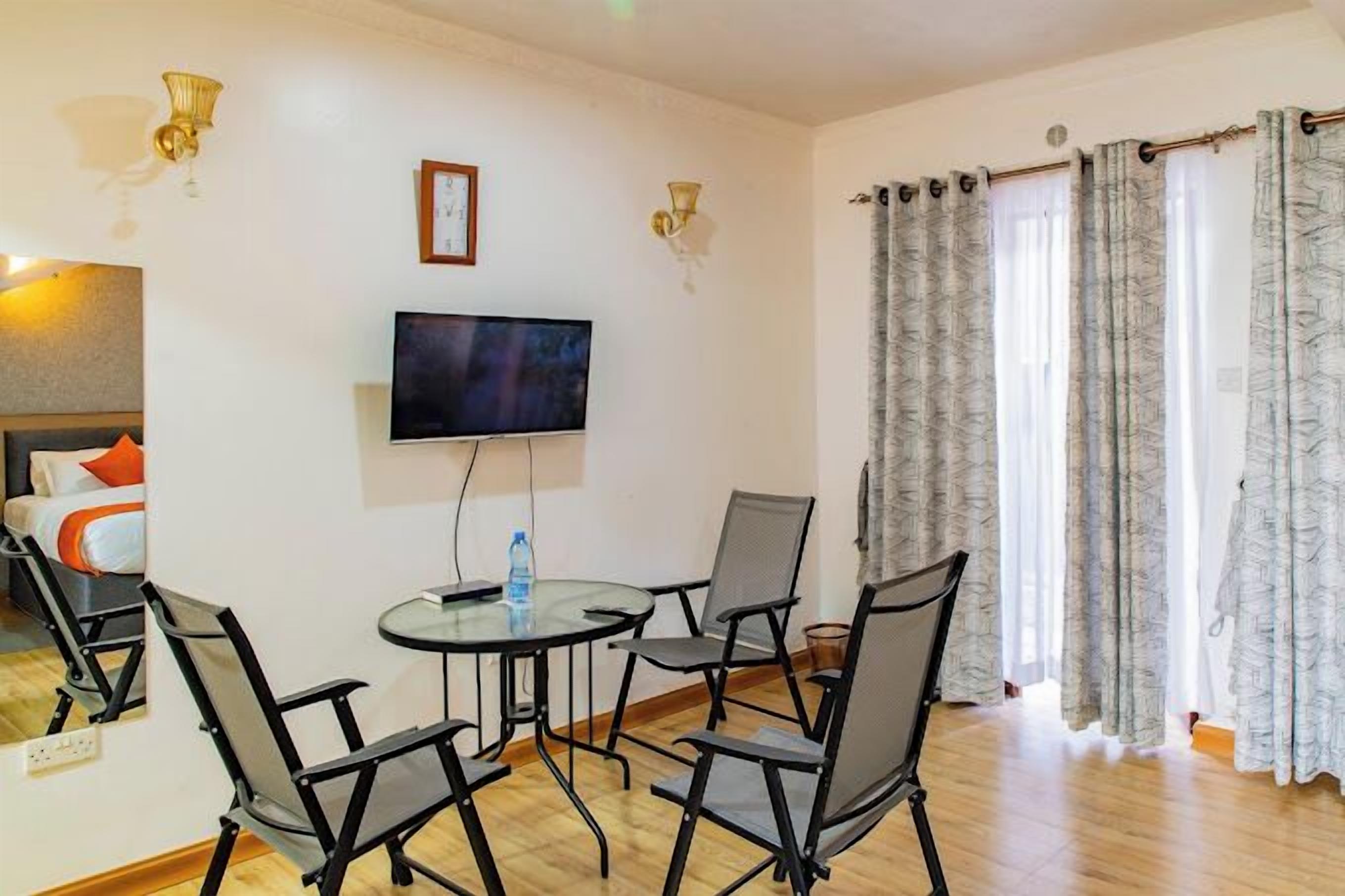 Hotellihuone, jossa on sänky, televisio, pyöreä pöytä tuoleineen ja ikkuna, jossa on verhot.
