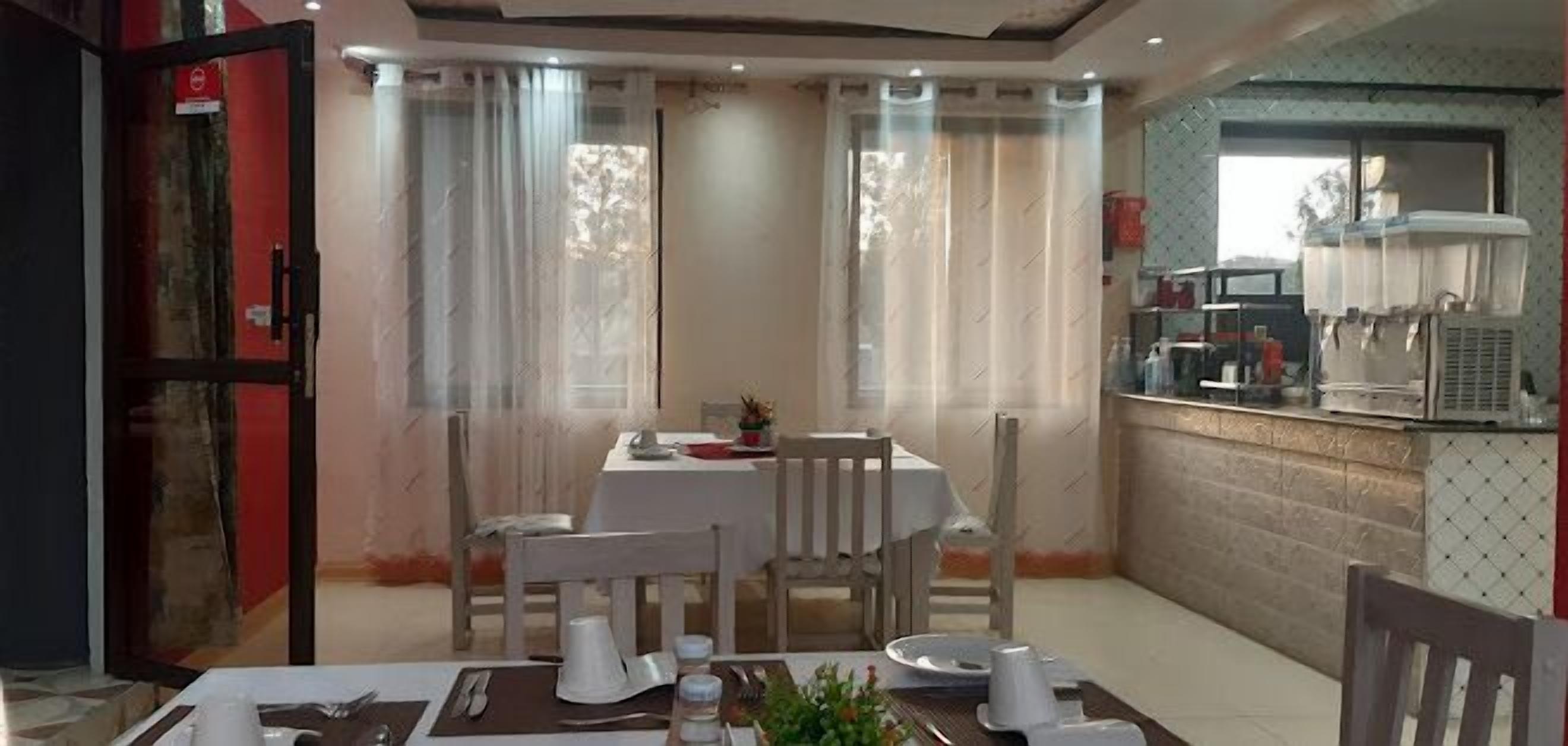 Un coin repas avec une table dressée pour deux personnes, un coin cuisine équipé d’un four à micro-ondes et d’un réfrigérateur, et une fenêtre agrémentée de rideaux légers.