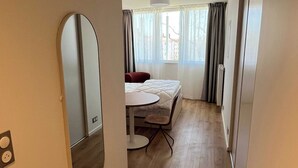A narrow hallway with a bed, a table, and a chair in a room with wooden flooring and a large mirror on the wall. - Comfort Studio in a Premium Residence- Sport/cinema (Huningue)