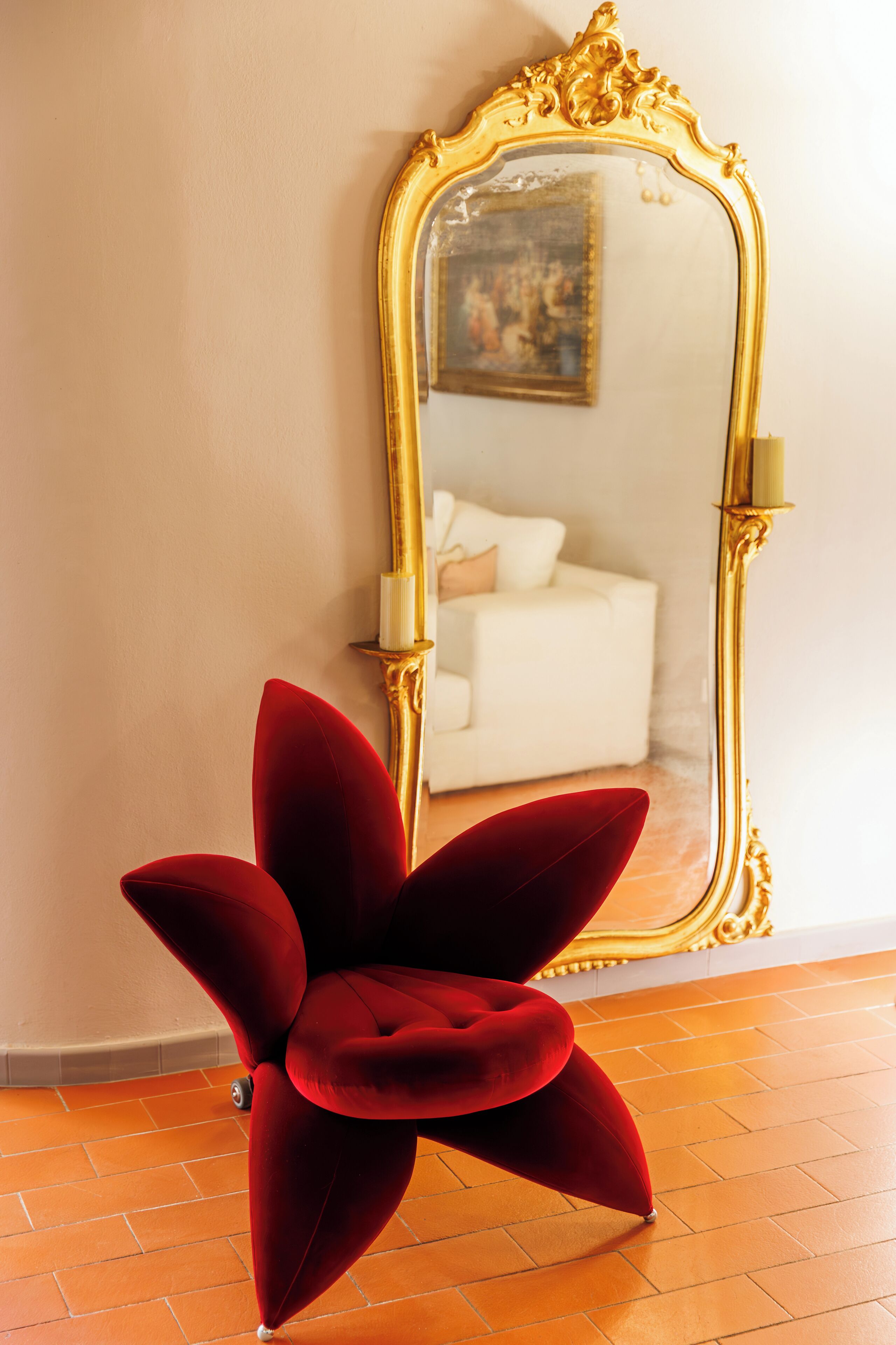 Un espejo con marco dorado, un sillón de terciopelo rojo y un sofá blanco al fondo.