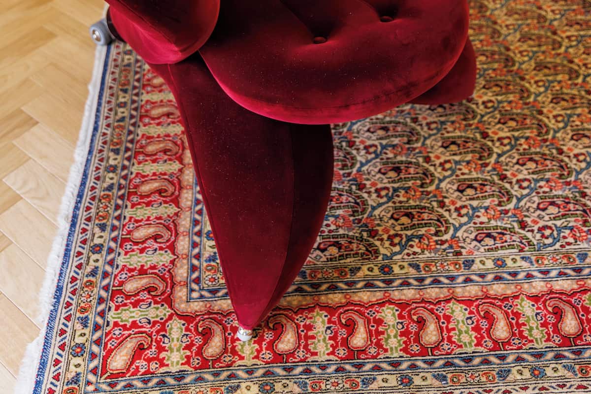 Un fauteuil en velours rouge, orné d’un grand nœud décoratif, se trouve dans une chambre d’hôtel. On y aperçoit également un tapis à motifs et un lit recouvert de linge de lit blanc.