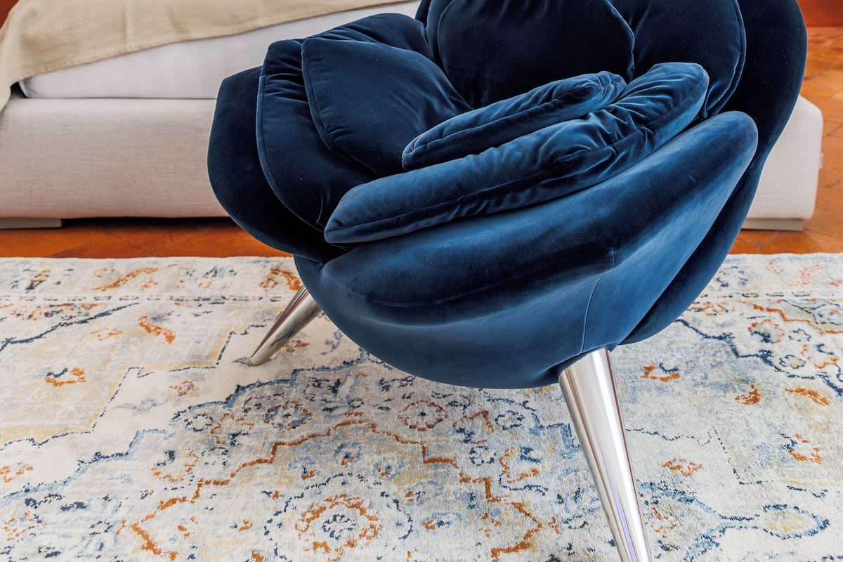 Une chambre moderne avec un fauteuil en velours bleu, un lit agrémenté d’oreillers jaunes et blancs, et un élément décoratif mural.
