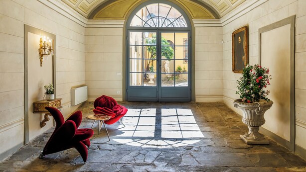 A hallway with a large arched doorway, a red chair, a small table, a wall-mounted light fixture, and a potted plant. - PALAZZO DELLA BIANCA- Royal Experience (Pisa)
