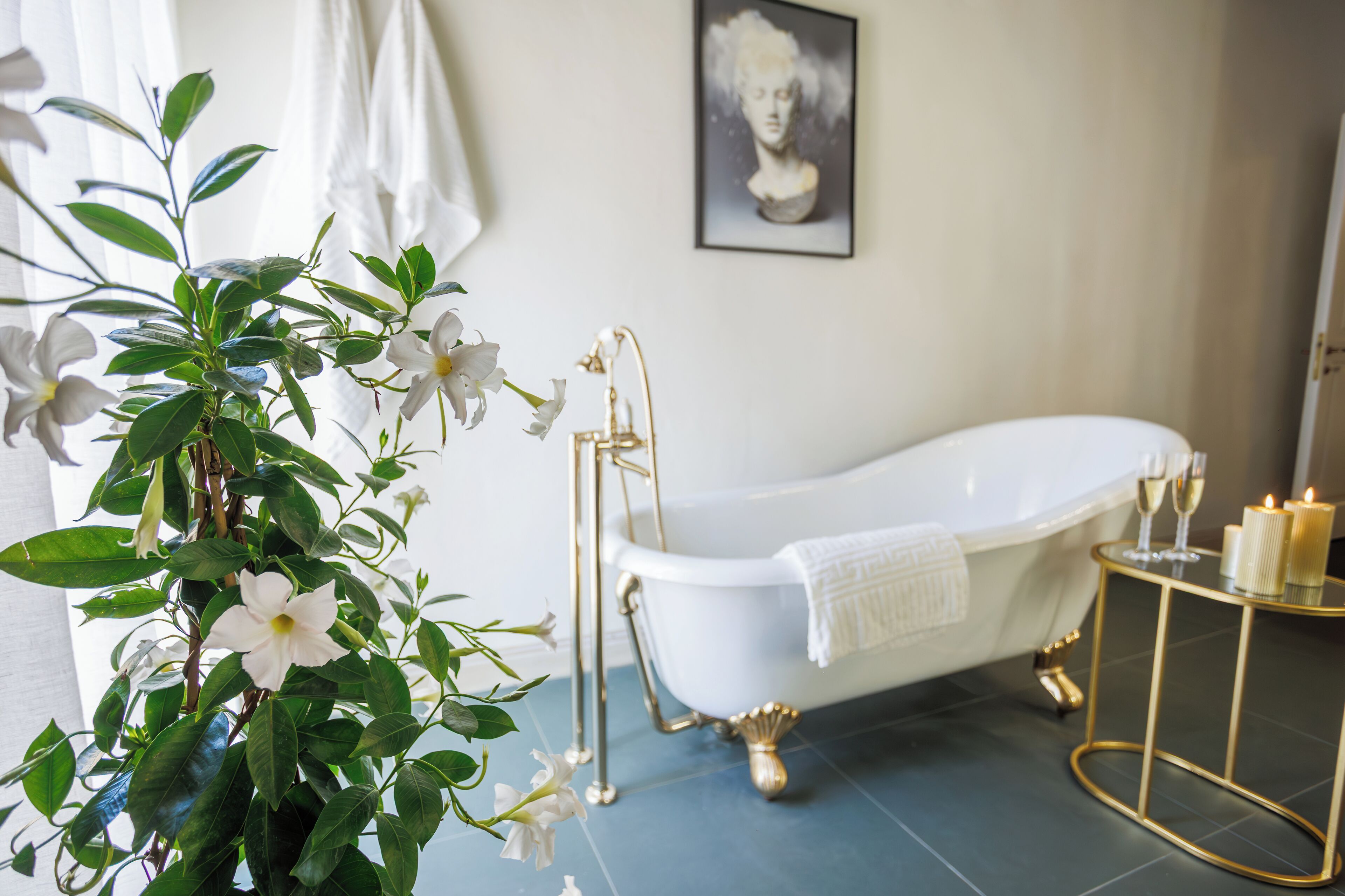 Een badkamer met een witte badkuip, een gouden badkuipstandaard, een gouden bijzettafeltje met kaarsen, en een lijstwerk met een kunstwerk aan de muur.