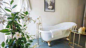 Een badkamer met een witte badkuip, een gouden badkuipstandaard, een gouden bijzettafeltje met kaarsen, en een lijstwerk met een kunstwerk aan de muur.