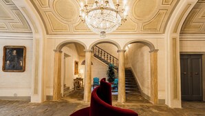A grand hallway with a chandelier, columns, a staircase, and a red chair. - PALAZZO DELLA BIANCA- Royal Experience (Pisa)