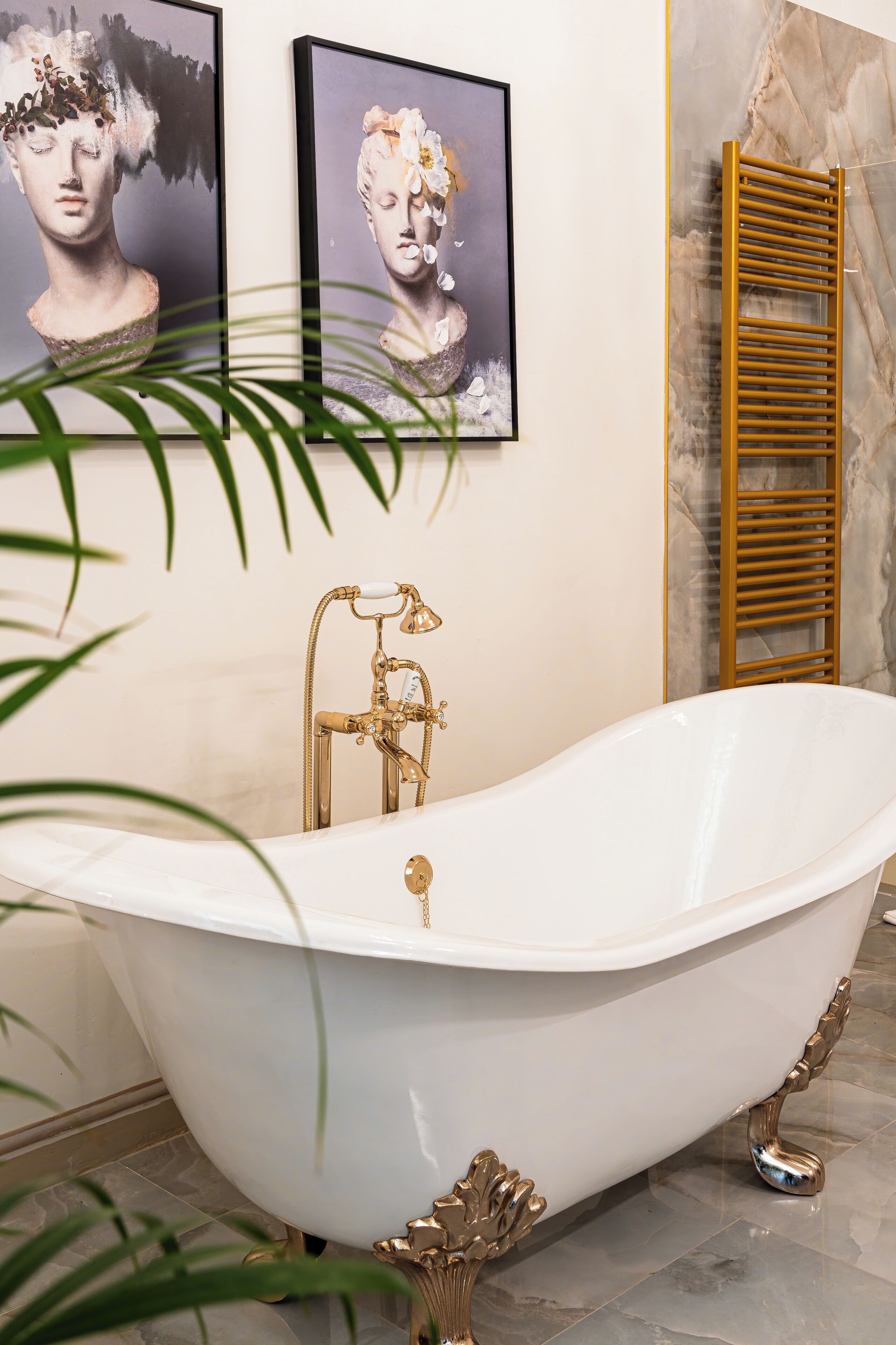 A white freestanding bathtub with gold fixtures, flanked by framed artwork and a wooden towel rack.