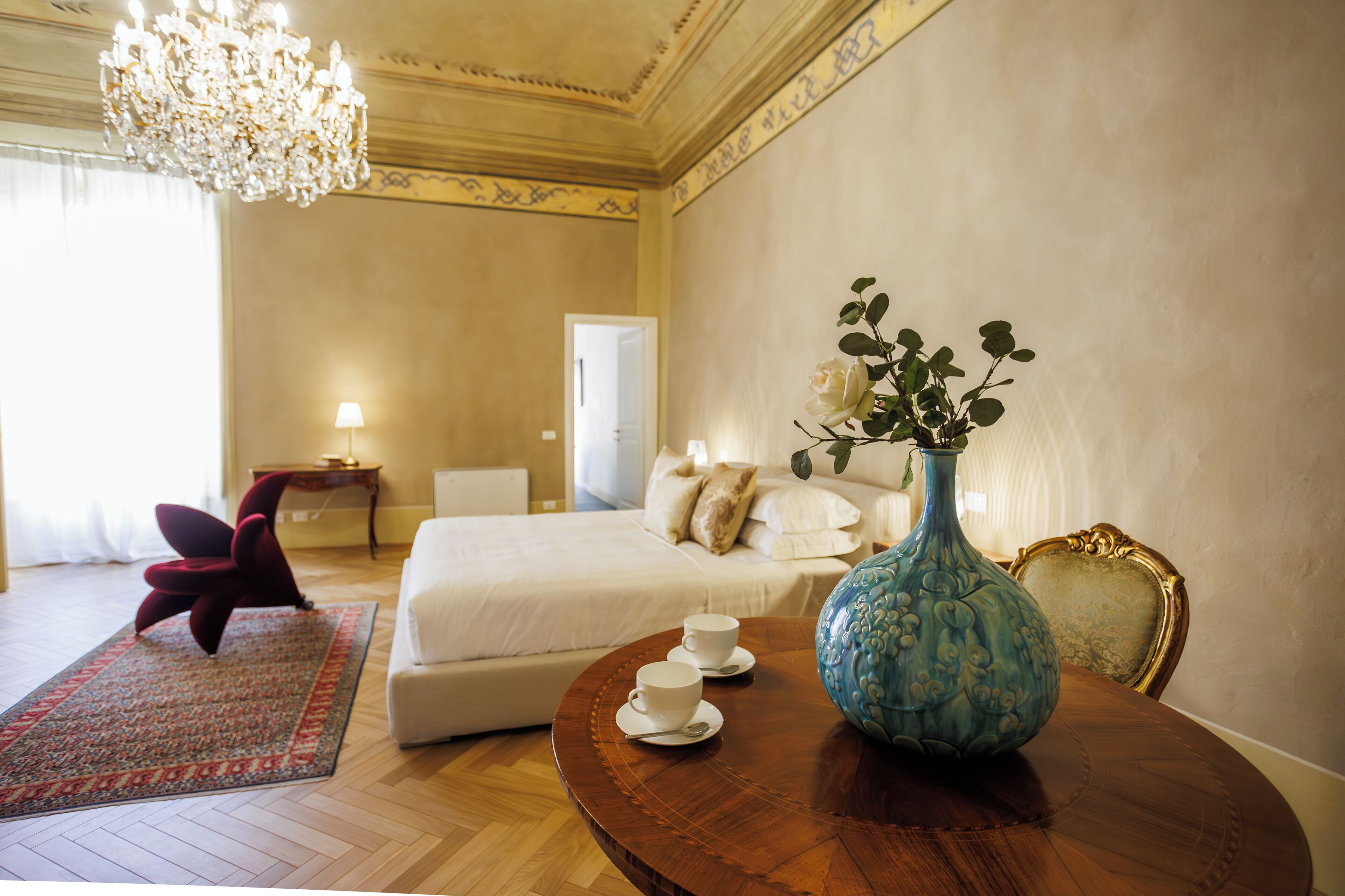 A hotel room with a bed, a round wooden table with a vase and cups, a red chair, and a chandelier.