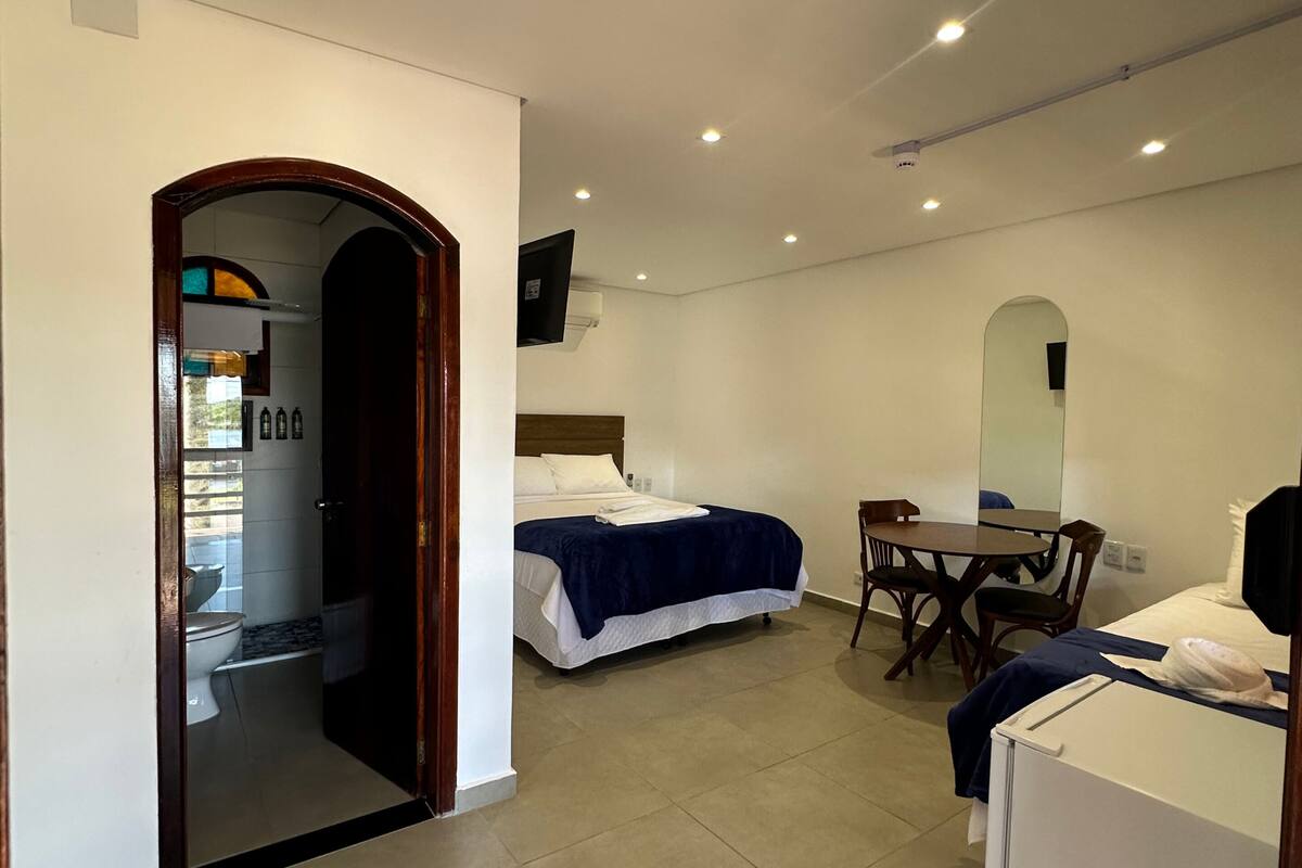 Habitación de hotel con dos camas, un escritorio, una silla, una mesita, un espejo y un baño visible a través de un arco.