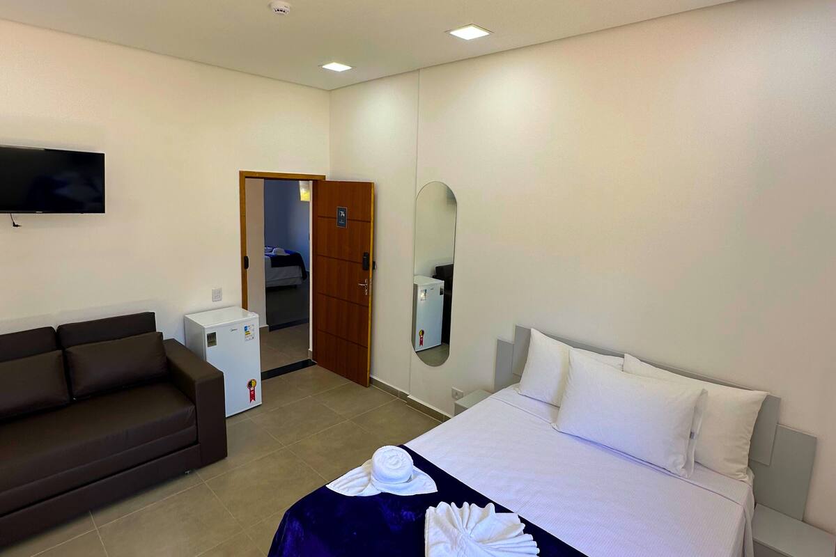 Habitación de hotel con cama, sofá, televisión, mini refrigerador y espejo.