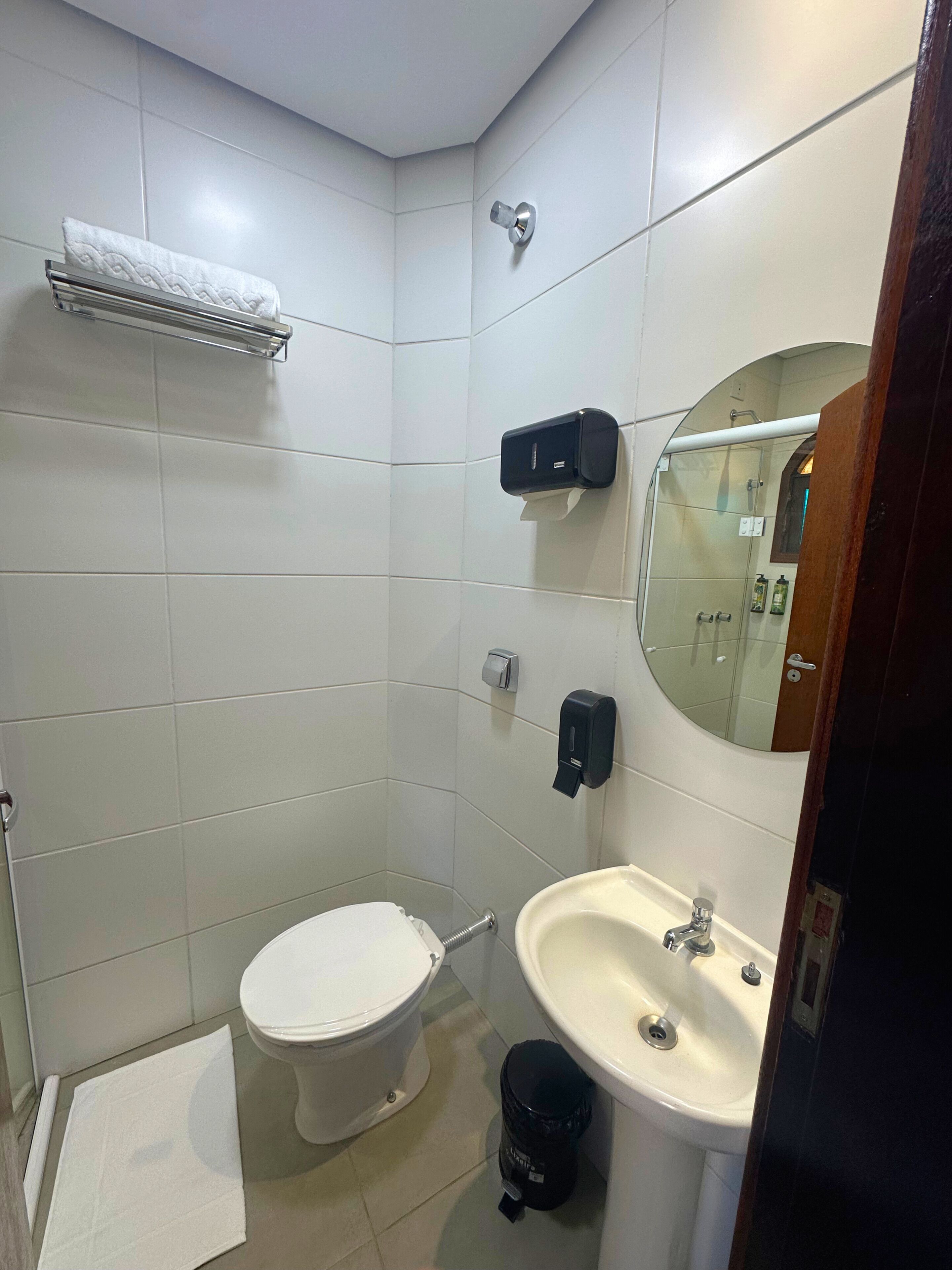 A small, clean bathroom with white tiles, a toilet, a sink, a mirror, and a towel rack.