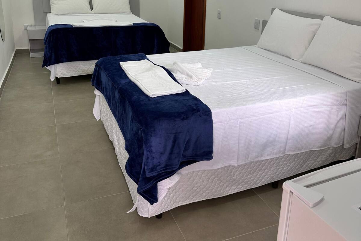 Habitación de hotel con dos camas, cada una con ropa blanca y una manta azul. El piso es de baldosas, la puerta de madera, y hay una mesita de noche con una lámpara.