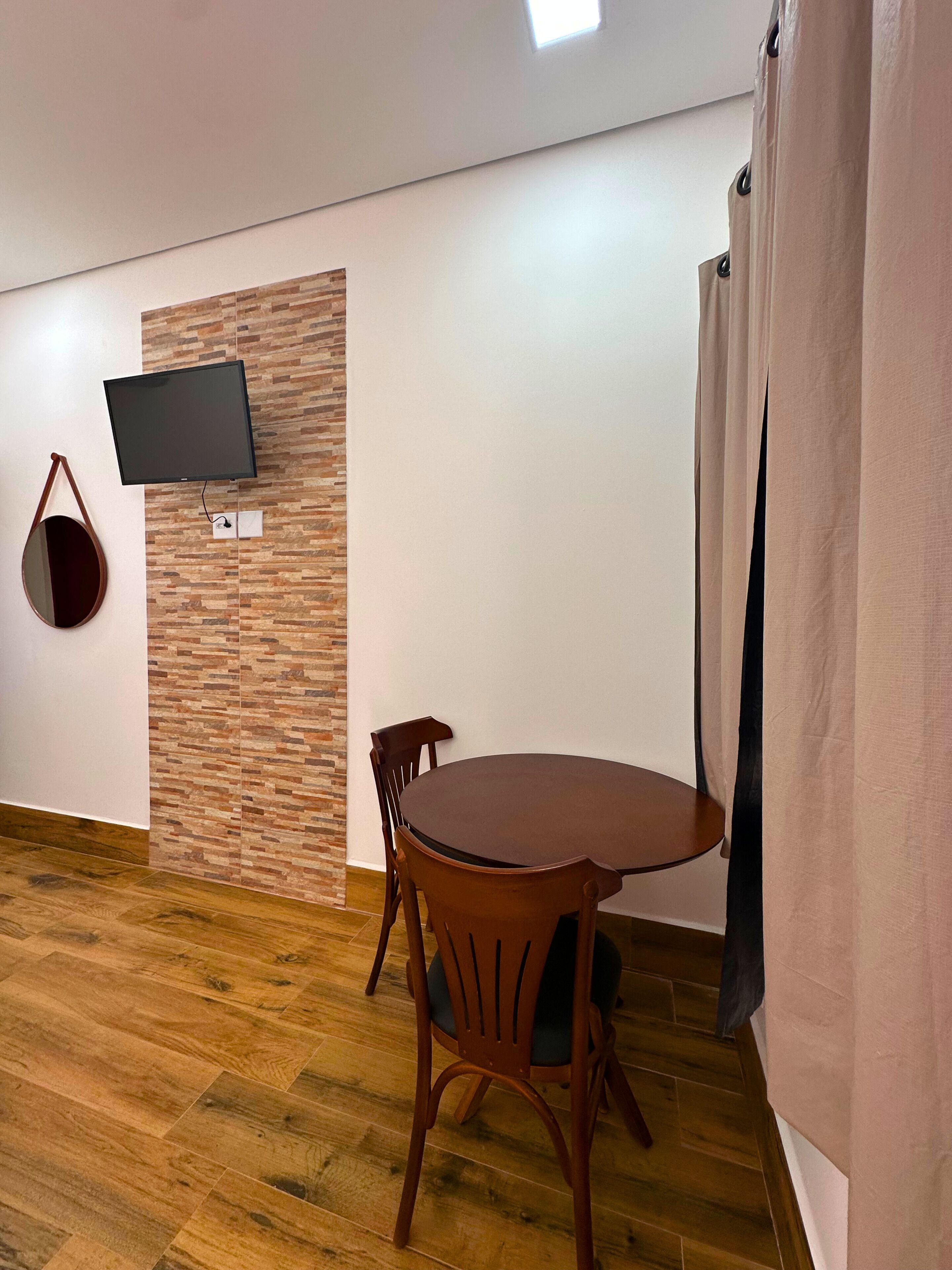 Una habitación con una mesa redonda de madera, dos sillas de madera, un televisor de pantalla plana montado en una pared con diseño de ladrillos y un bolso colgando.