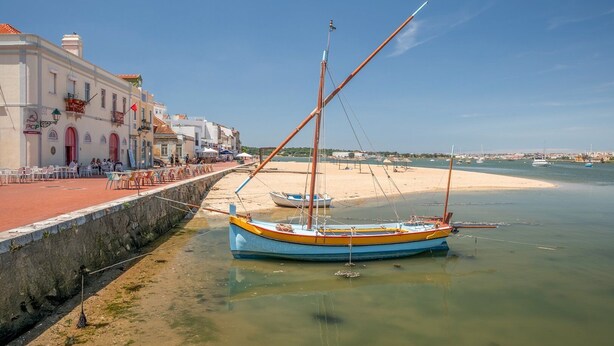 A coastal scene with a boat docked near a sandy beach, a stone wall, and a building with outdoor seating. - GuestReady - Beautiful stay in Seixal (Seixal)