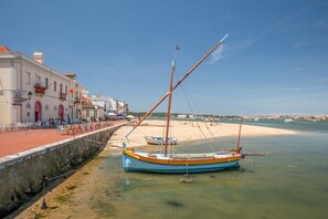 A coastal scene with a boat docked near a sandy beach, a stone wall, and a building with outdoor seating.