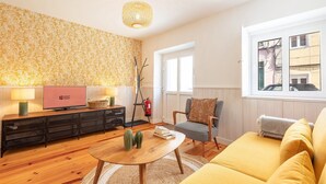 A living room with a wooden floor, a yellow sofa, a television, a patterned wallpaper, and a window with a view of buildings. - GuestReady - Beautiful stay in Seixal (Seixal)